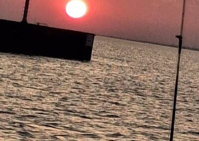 A vibrant orange and pink sunset glows over slightly choppy water, with a dark structure topped by a navigational marker on the left and a fishing rod visible in the right foreground - foxyladycharterservice.com