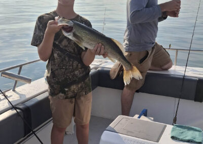 A young boy standing on a boat proudly holds up a large fish he caught, while a man prepares fishing gear in the background against a calm body of water - foxyladycharterservice.com