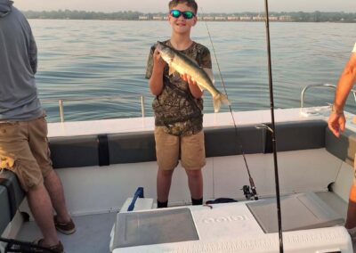 A young boy standing on a boat smiles while holding up a freshly caught fish, with fishing gear, a cooler, and open water in the background - foxyladycharterservice.com