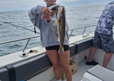 A smiling woman stands on a fishing boat holding up a freshly caught walleye fish using a plastic lip gripper tool, with open water and a cloudy sky in the background