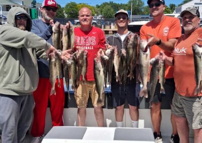 Six men stand behind a white cooler on a boat deck under a clear blue sky, smiling and proudly holding up a large catch of fish - foxyladycharterservice.com
